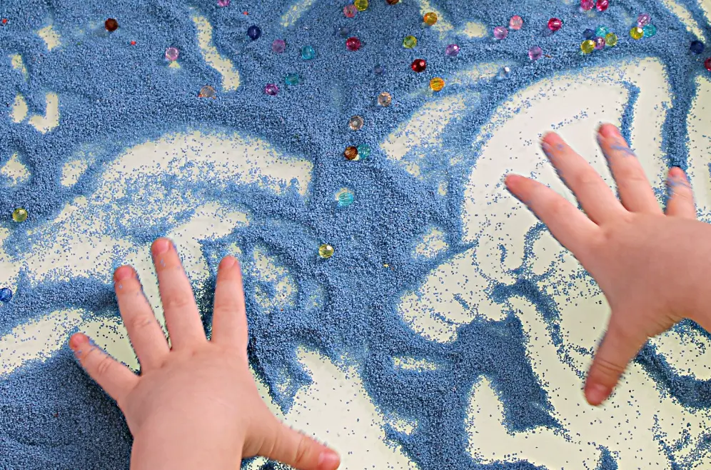 Hands of a child playing with blue sand with gems in it.