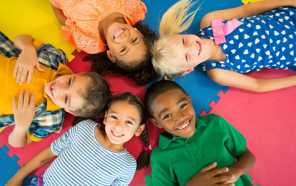 A group of kids laying on a colored mat with their heads touching and smiling.